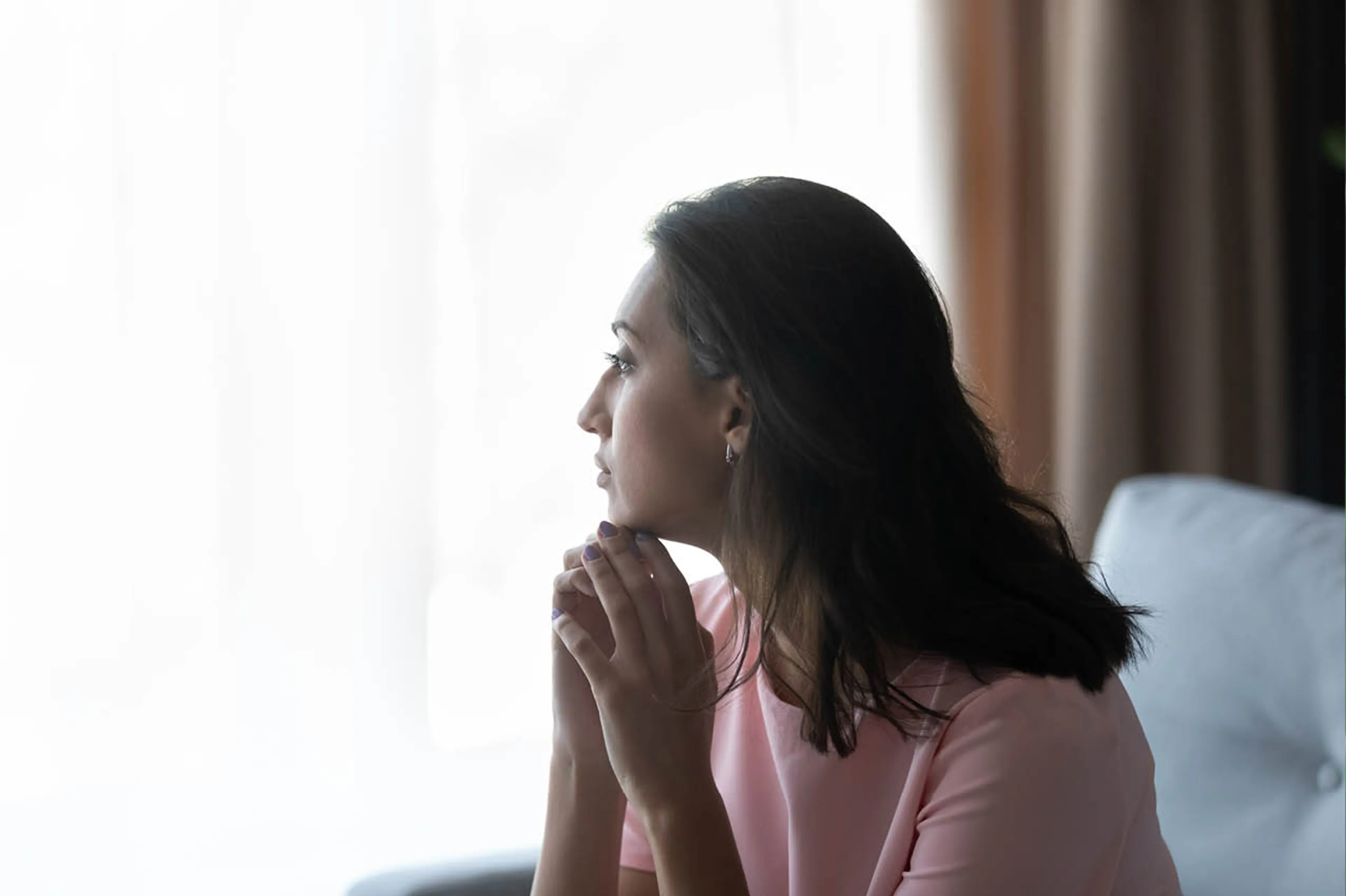 Vrouw met bruin haar, kijkt uitdrukkingsloos uit het raam en zit op feuteuil